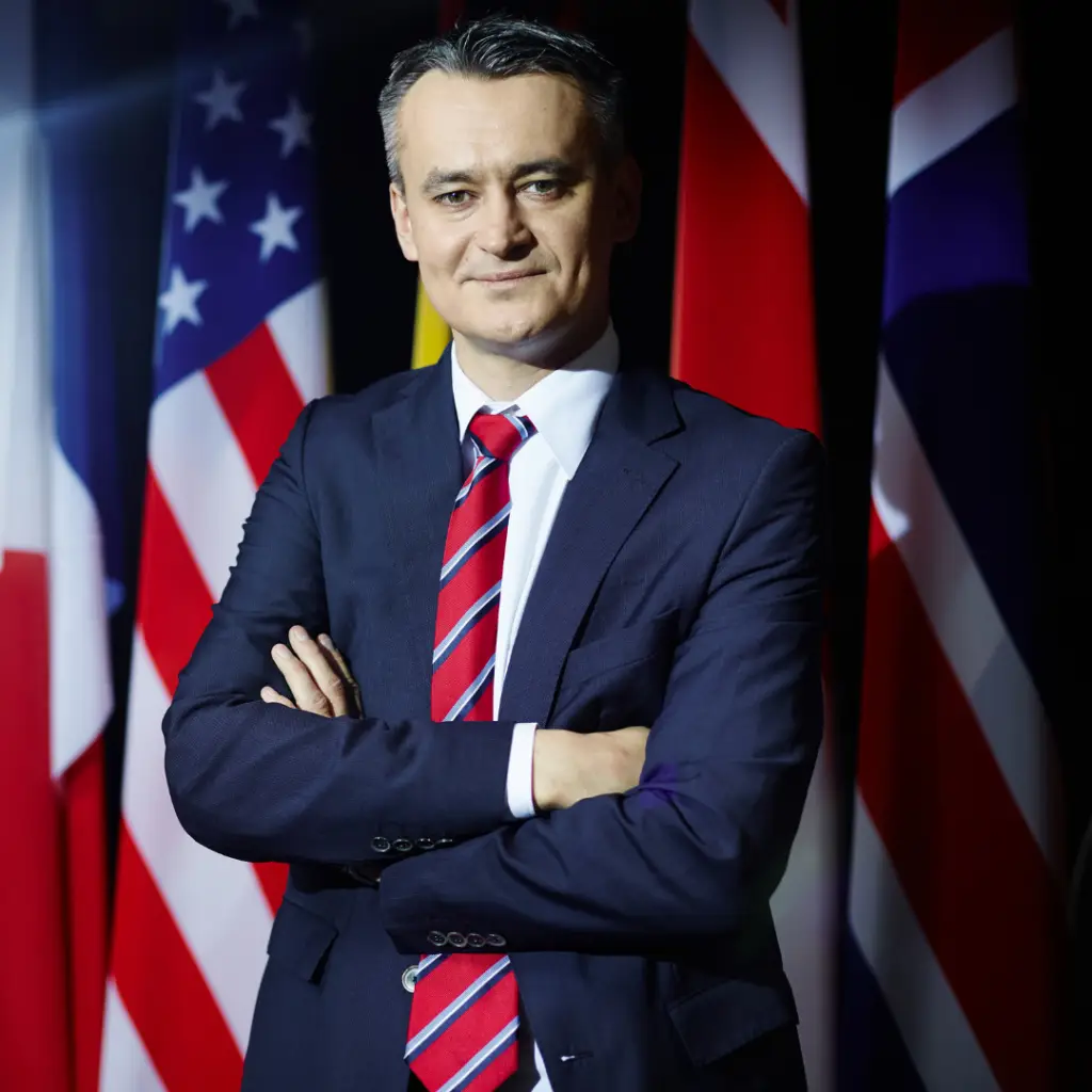 Politician in blue suit and red tie and arms crossed in front of the USA, British and French flags